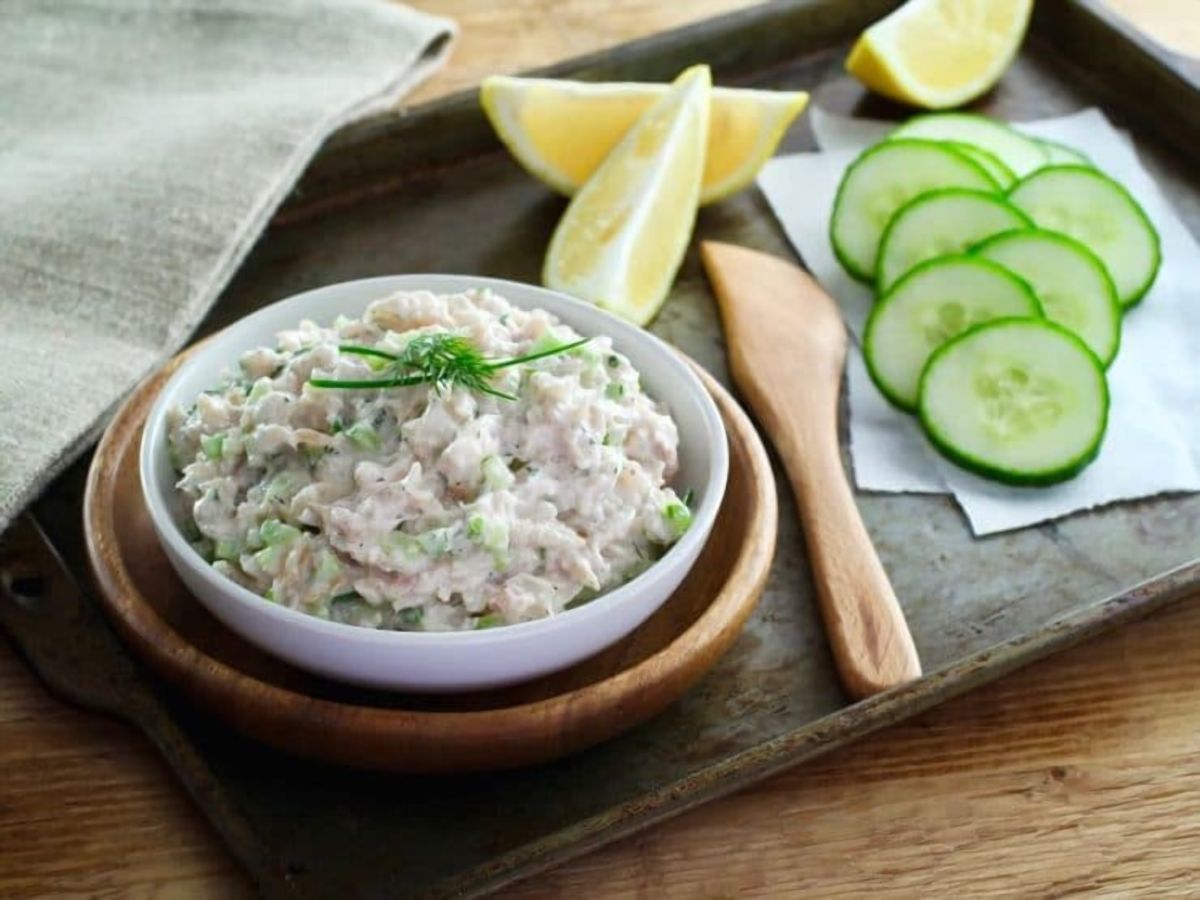 Horizontal shot of whitefish salad in a white bowl, nestled inside a larger brown bowl with a brown spatula. Sliced lemon and cucumber on the right, placed atop a brown tray.