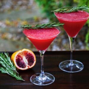 Horizontal shot of two martini glasses filled with a bright red blood orange cocktail. Both are garnished with a sprig of fresh rosemary. A sliced blood orange and more rosemary sit off to the left.