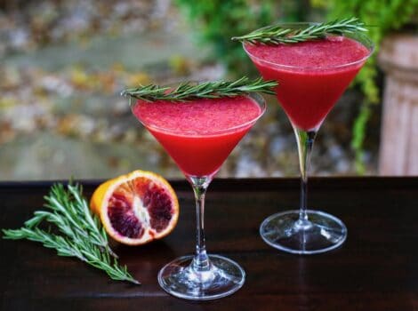 Horizontal shot of two martini glasses filled with a bright red blood orange cocktail. Both are garnished with a sprig of fresh rosemary. A sliced blood orange and more rosemary sit off to the left.