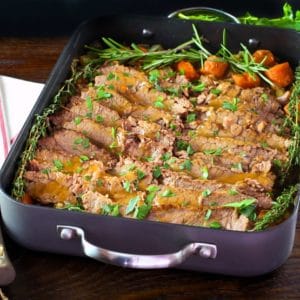 Roasting pan with cooked sliced brisket and carrots topped with fresh herbs, fresh parsley and cloth napkin on the side.