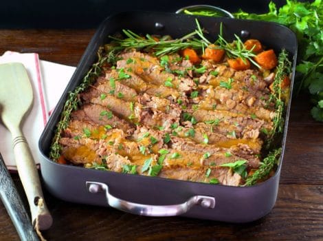 Roasting pan with cooked sliced brisket and carrots topped with fresh herbs, fresh parsley and cloth napkin on the side.
