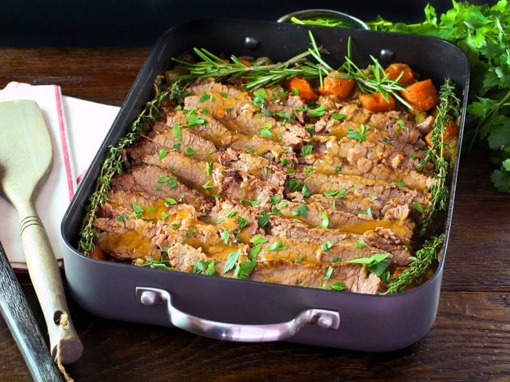 Roasting pan with cooked sliced brisket and carrots topped with fresh herbs, fresh parsley and cloth napkin on the side.