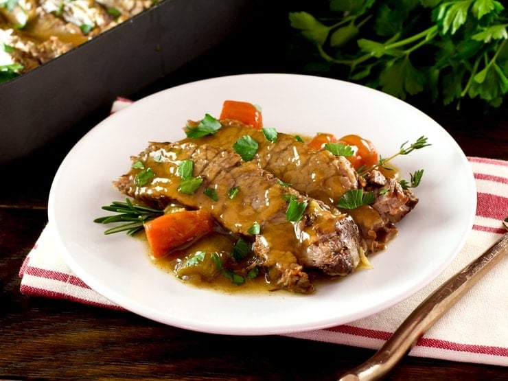 White plate with tender slices of savory herb braised brisket and carrots, topped with fresh herbs. Napkin, fork and fresh herbs in background with roasting pan of meat.