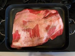 Uncooked brisket coated with salt and pepper in roasting pan on stovetop.