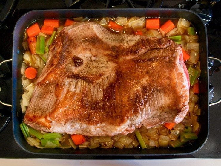Browned brisket on top of onions, carrots and celery chopped in roasting pan on stovetop.