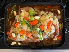 Browned brisket topped with garlic cloves, fresh herbs and vegetables on top of onions, carrots and celery chopped in roasting pan on stovetop.
