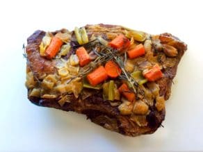 Cooked brisket topped with cooked vegetables and herbs on white cutting board.