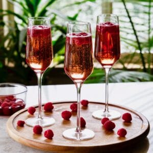 Horizontal shot of three kir royale drinks with Moscato. Cocktails in champagne flutes sitting on a round wooden serving tray. Fresh raspberries are scattered across the tray and a dish of fresh raspberries sits off to the left.