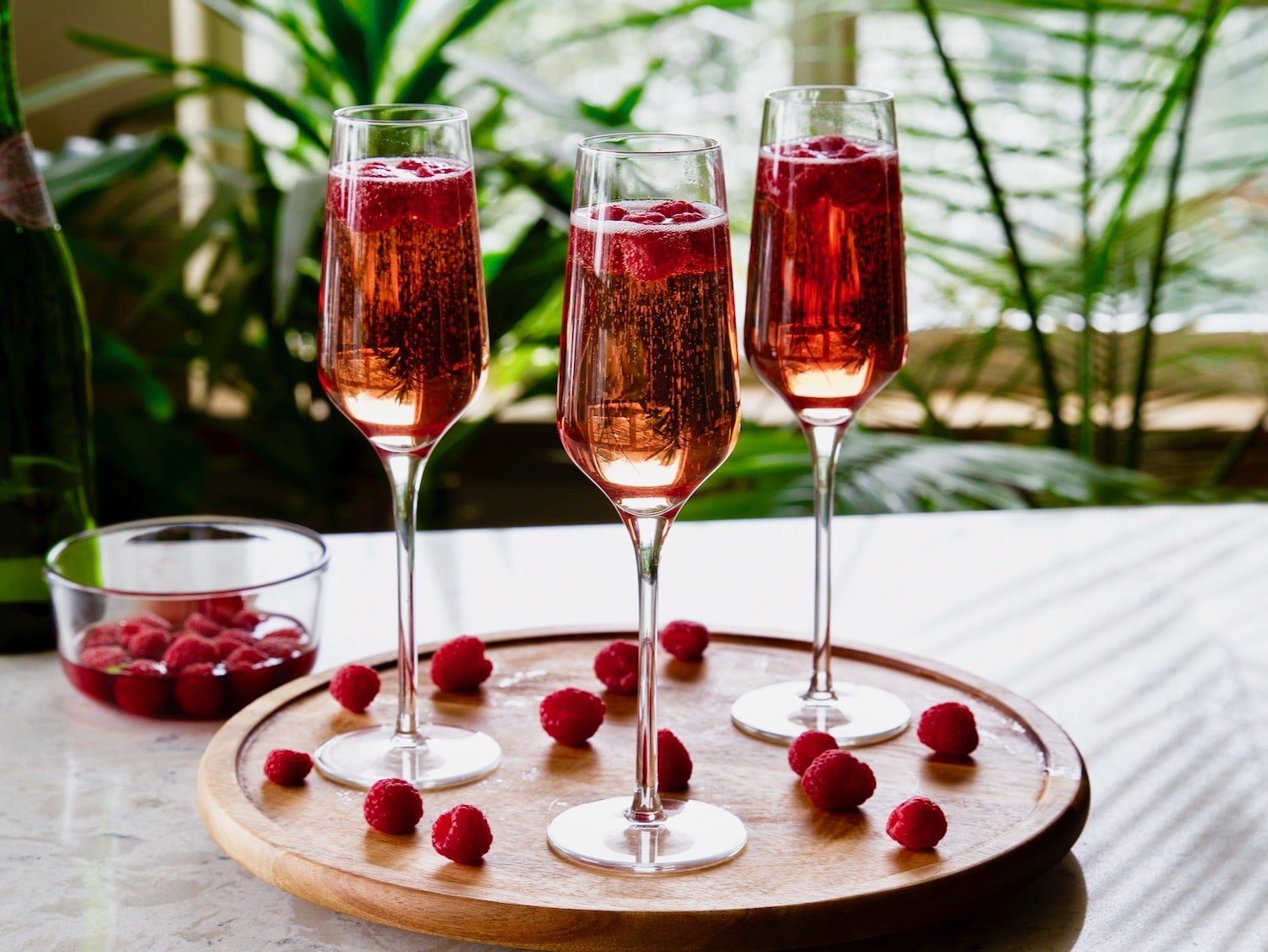 Horizontal shot of three kir royale drinks with Moscato. Cocktails in champagne flutes sitting on a round wooden serving tray. Fresh raspberries are scattered across the tray and a dish of fresh raspberries sits off to the left.