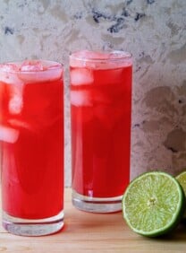 Horizontal shot of two red cocktails in tall glasses filled with ice. A sliced lime sits next to the glass on the right.