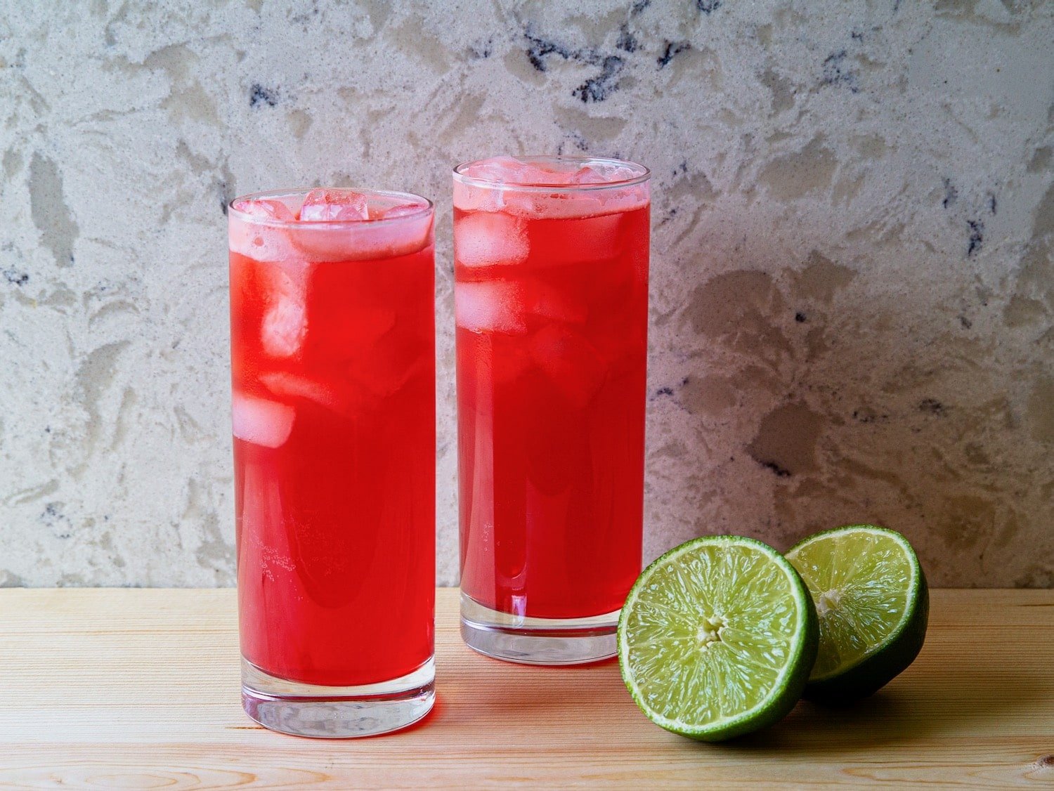 Horizontal shot of two red cocktails in tall glasses filled with ice. A sliced lime sits next to the glass on the right.