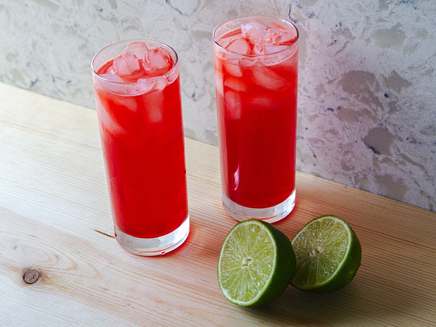 Horizontal angled shot of two red cocktails in tall glasses filled with ice. A sliced lime sits next to the glass on the right.