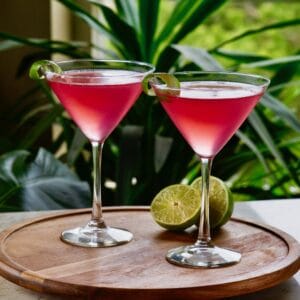 Horizontal shot of two red cosmos - cranberry vodka cosmopolitan cocktails in martini glasses on top of a round wooden serving tray. A sliced lime sits behind the glass on the right.