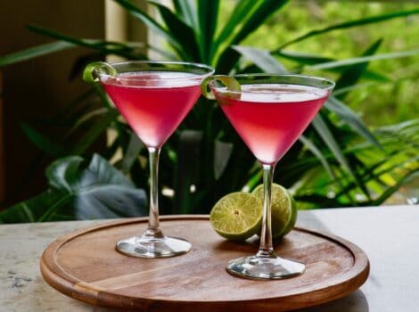 Horizontal shot of two red cosmos - cranberry vodka cosmopolitan cocktails in martini glasses on top of a round wooden serving tray. A sliced lime sits behind the glass on the right.