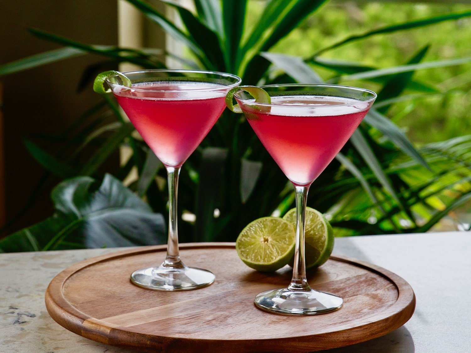 Horizontal shot of two red cosmopolitan cocktails in martini glasses on top of a round wooden serving tray. A sliced lime sits behind the glass on the right.