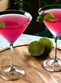 Horizontal angled shot of two cosmos - red cranberry vodka cosmopolitan cocktails in martini glasses on top of a round wooden serving tray. A sliced lime sits behind the glass on the right.