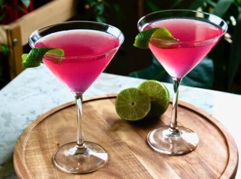 Horizontal angled shot of two cosmos - red cranberry vodka cosmopolitan cocktails in martini glasses on top of a round wooden serving tray. A sliced lime sits behind the glass on the right.