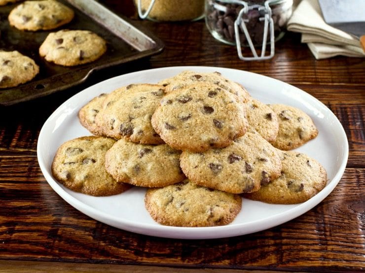 Close up of Passover matzo chocolate chip cookies on white plate on wooden background with jar of chocolate chips, jar of brown sugar, napkins and baking tray in background.