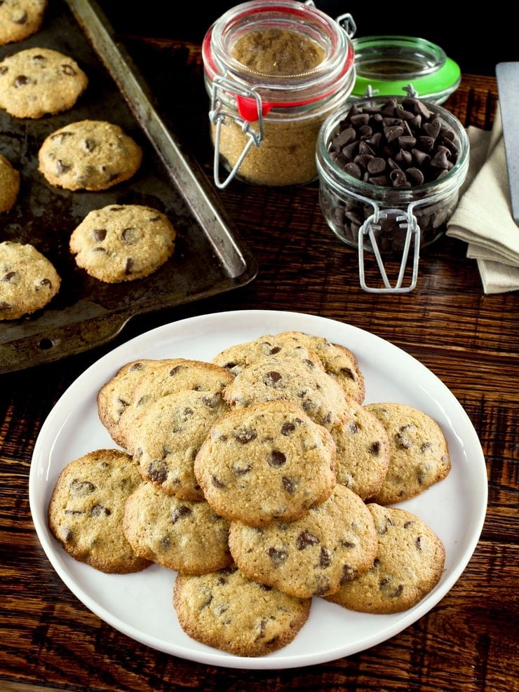 Overhead vertical shot of Passover matzo chocolate chip cookies on white plate on wooden background with jar of chocolate chips, jar of brown sugar, napkins and baking tray in background.