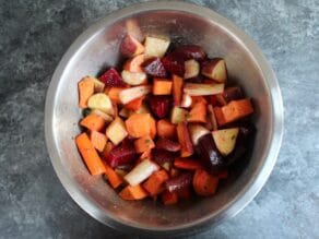 Overhead shot of raw chunks of vegetables - beets, parsnips, potatoes, carrots - in a mixing bowl on a concrete background.