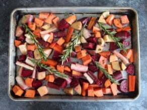 Large rimmed baking sheet with root vegetable chunks placed evenly across, topped with 5 sprigs of fresh rosemary.