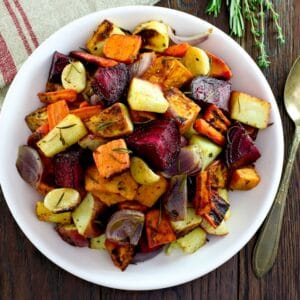 Overhead shot of oven roasted root vegetables with beets, carrots, potatoes, and parsnip. Spoon and fresh herbs on the side, cloth napkin above.