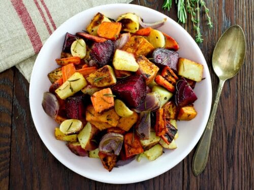 Overhead shot of oven roasted root vegetables with beets, carrots, potatoes, and parsnip. Spoon and fresh herbs on the side, cloth napkin above.