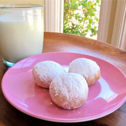 Kourabiedes Greek butter cookies on a pink circular plate, paired elegantly with a glass of milk, all atop a rustic brown table.