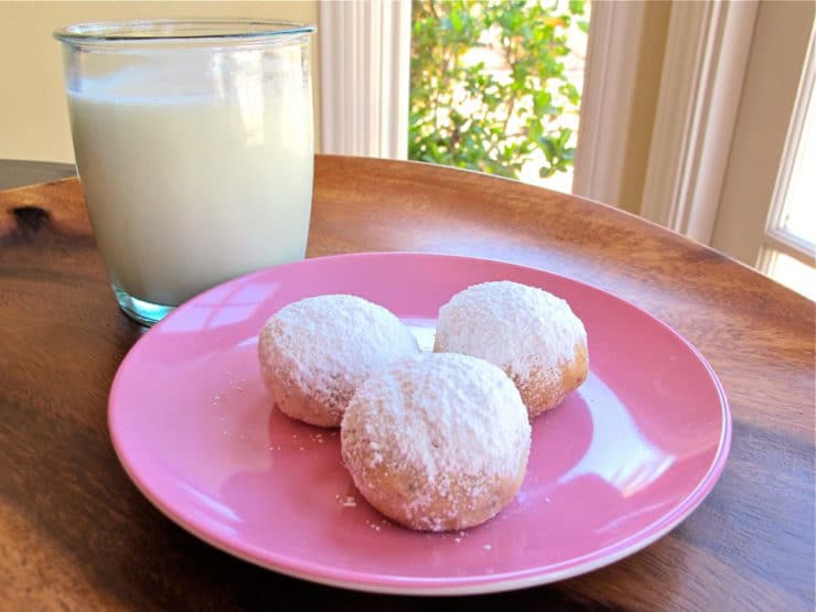 Kourabiedes Greek butter cookies on a pink circular plate, paired elegantly with a glass of milk, all atop a rustic brown table.