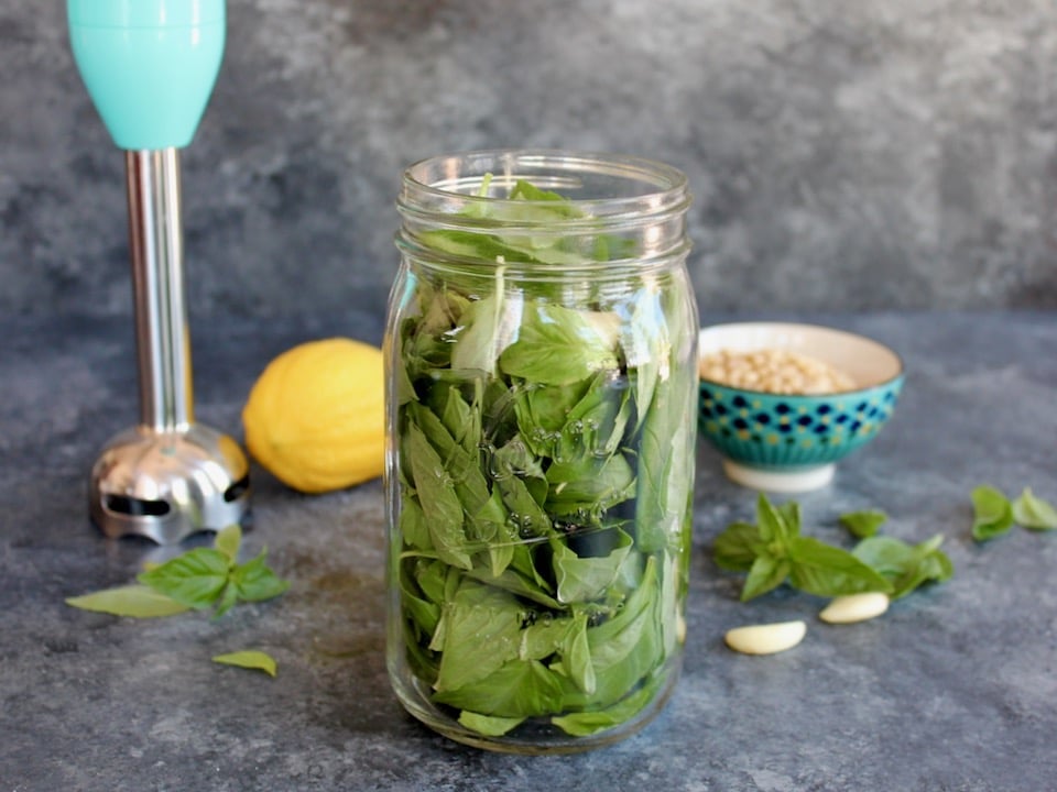 Vertical shot of a large mason jar filled with fresh basil leaves sitting in front of a small decorative bowl, a lemon, and an immersion blender.