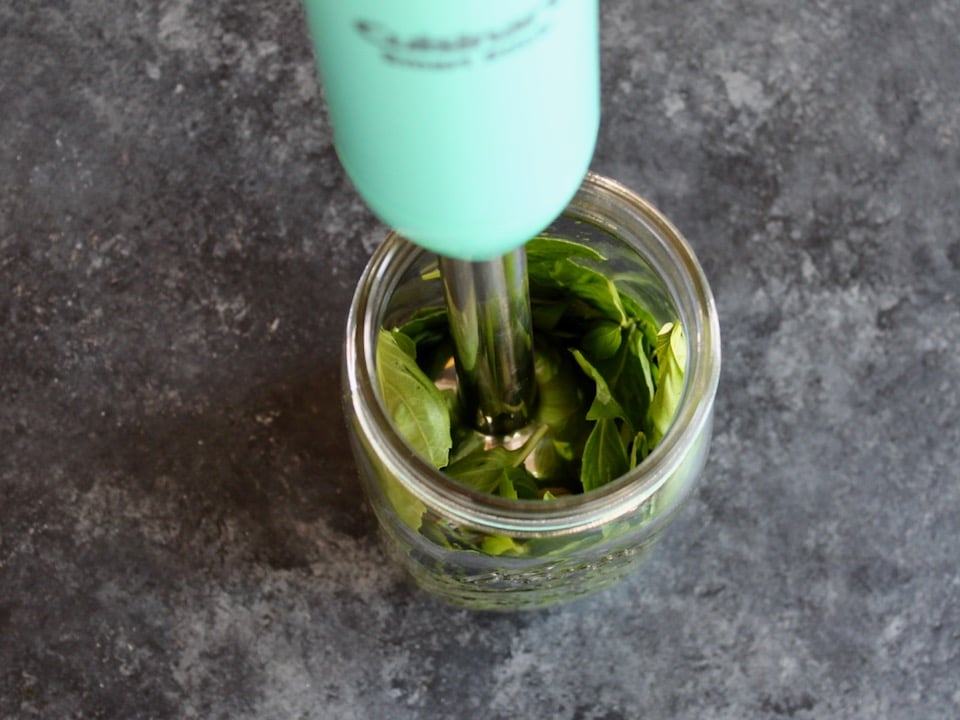 Overhead shot of an immersion blender blending fresh basil leaves inside of a glass mason jar.