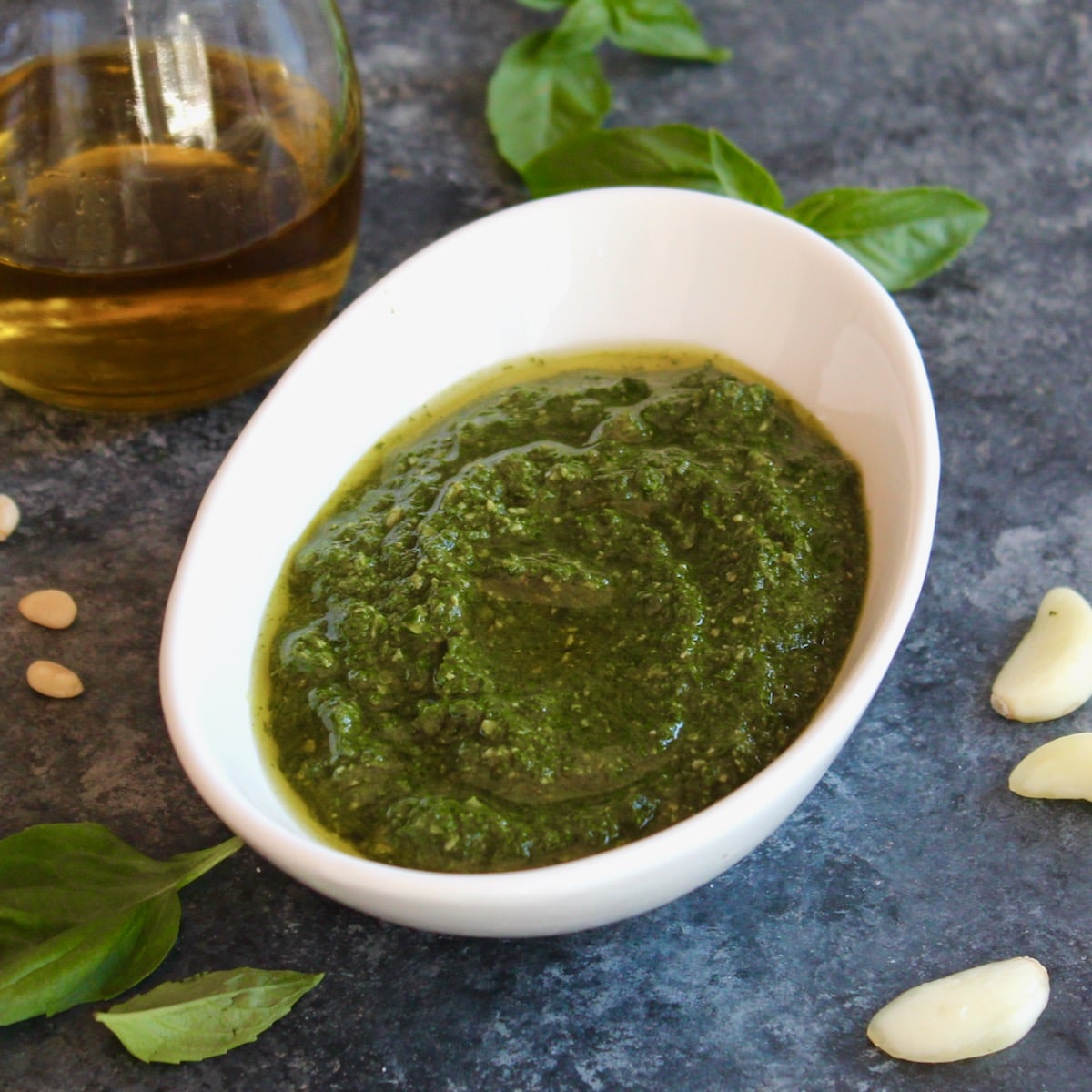 Square shot of a decorative white bowl filled with fresh basil pesto, the area around the bowl is sprinkled with fresh basil leaves and garlic cloves. A jug of olive oil sits off to the left.