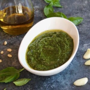 Horizontal shot of a decorative white bowl filled with fresh basil pesto, the area around the bowl is sprinkled with fresh basil leaves and garlic cloves. A jug of olive oil sits off to the left.