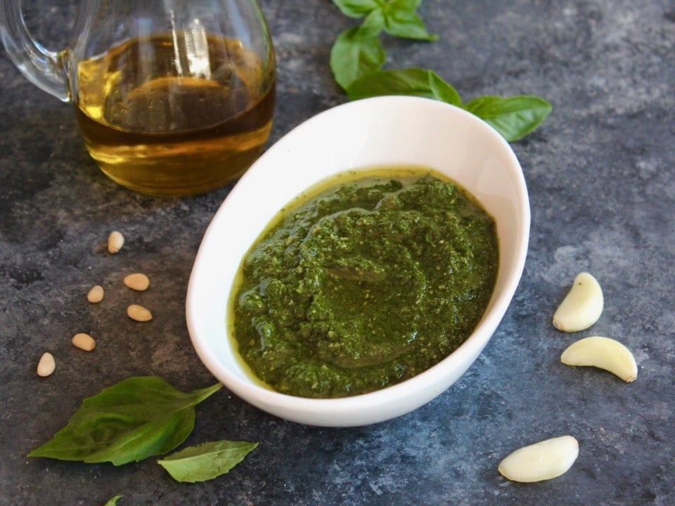 Horizontal shot of a decorative white bowl filled with fresh basil pesto, the area around the bowl is sprinkled with fresh basil leaves and garlic cloves. A jug of olive oil sits off to the left.