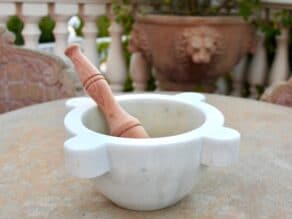 Traditional Italian marble mortar and wooden pestle on a stone table outside, chair and planter with lion's head in background, balustrades in background.