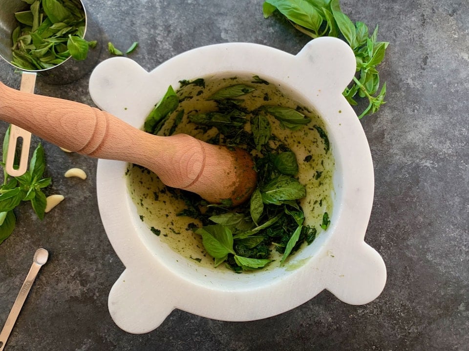 Italian marble mortar with wooden pestle, basil leaves half-crushed, on countertop with basil, garlic cloves and a spoon of salt laying beside it.