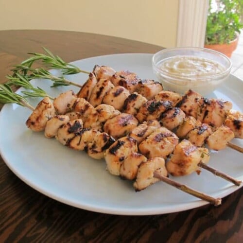 A horizontal shot of Rosemary Lemon Chicken Skewers neatly served on a white round plate with a sauce on a small bowl beside