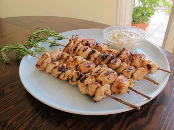 A horizontal shot of Rosemary Lemon Chicken Skewers neatly served on a white round plate with a sauce on a small bowl beside