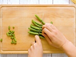 Hand slicing green beans on a wooden cutting board, trimmings off to the side.