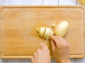 Hand cutting peeled potato into large chunks on a wooden cutting board.