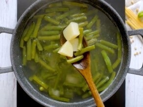 Wooden spoon scooping out cooked potatoes and green beans from a large pot of cooked vegetables in hot water on an induction cooktop.