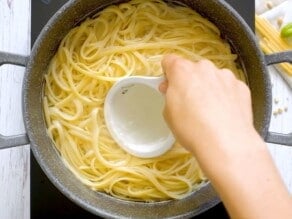 Hand scooping pasta water from a large pot of cooked pasta using a measuring cup.