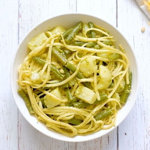 Overhead shot - bowl of trenette al pesto pasta with green beans and potatoes on a white wooden background, pine nuts and lemon on the side.