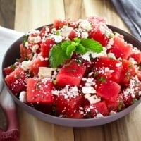 Beauty shot of watermelon salad with crumbled feta cheese and a mint garnish on a wooden cutting board with towels, napkin and spoon.