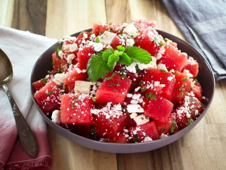Beauty shot of watermelon salad with crumbled feta cheese and a mint garnish on a wooden cutting board with towels, napkin and spoon.