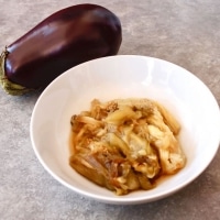 A whole eggplant beside a bowl of cooked eggplant on a grey countertop