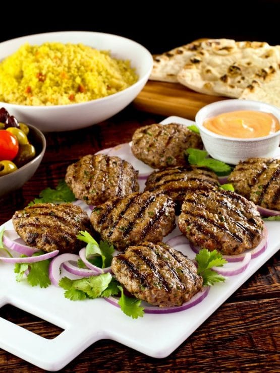 White platter of Middle Eastern Burgers char-grilled on a bed of purple onion rings and herbs, with spicy sriracha mayo sauce on the side, dish of yellow couscous, pita bread and olives in background.