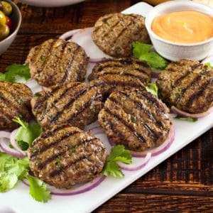 White platter of Middle Eastern Burgers char-grilled on a bed of purple onion rings and herbs, with spicy sriracha mayo sauce on the side, dish of olives in background.