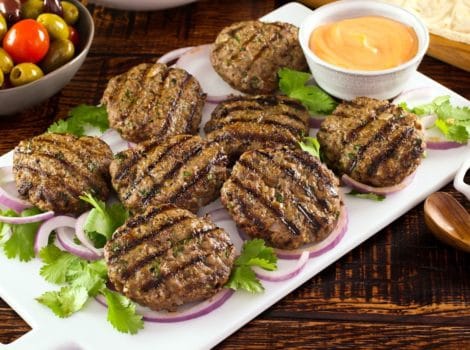 White platter of Middle Eastern Burgers char-grilled on a bed of purple onion rings and herbs, with spicy sriracha mayo sauce on the side, dish of olives in background.