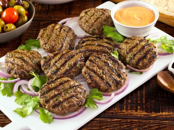 White platter of Middle Eastern Burgers char-grilled on a bed of purple onion rings and herbs, with spicy sriracha mayo sauce on the side, dish of olives in background.